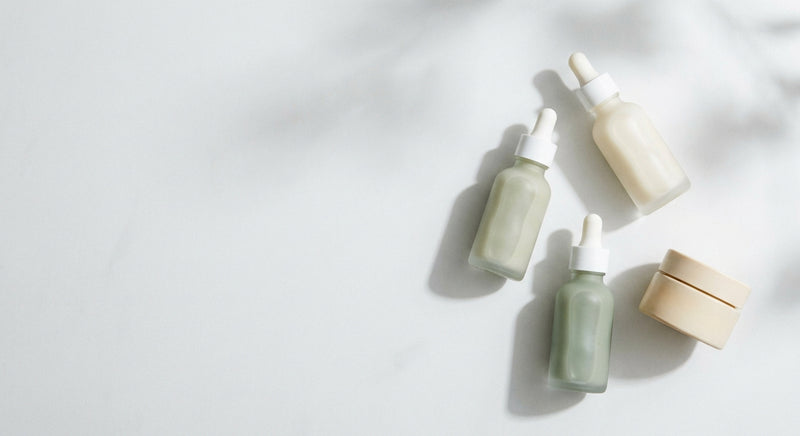 A minimalist flat-lay of a few clean-beauty skincare bottles and a ceramic jar on a white stone surface bathed in soft natural light.